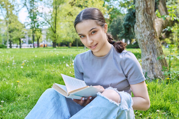 Teenage girl high school student reading book sitting on grass in park