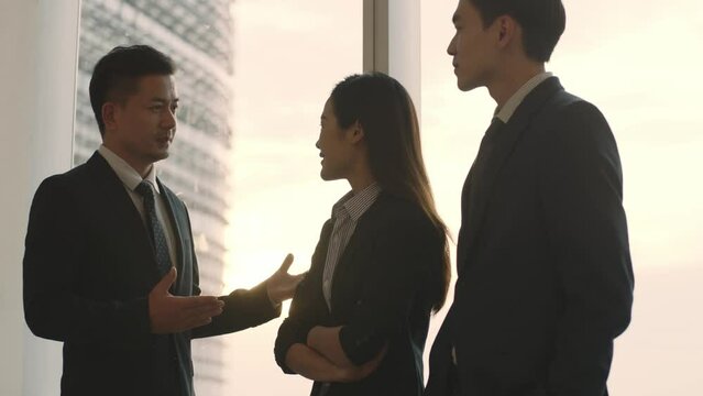 three asian business people colleagues standing and talking by the window in office at dusk