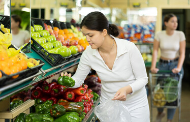 In vegetable department of supermarket,asian woman shopper thoughtfully chooses large fruits of Bulgarian pepper