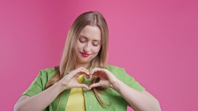 Smiling pretty woman showing sign of shape heart. Positive lady in pink studio