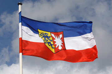 Flagge von Schleswig-Holstein mit Dienstwappen im Wind. Himmel mit Wolken. Die Flagge ist einmal um den Flaggenmast gewickelt.