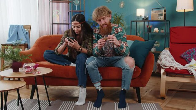 Family married hippie couple man woman using credit bank card and smartphone while transferring money, purchases online shopping, order food delivery at home. Husband and wife together on sofa in room