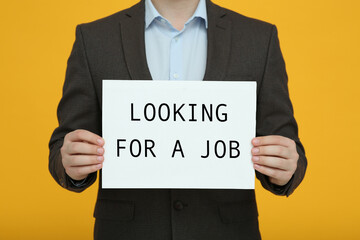 Unemployed man holding sign with phrase Looking For A Job on orange background, closeup