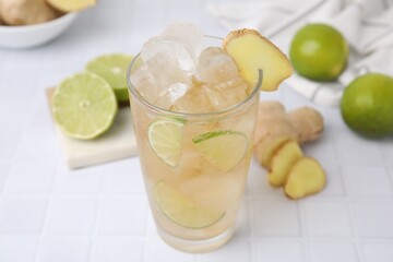 Glass of tasty ginger ale with ice cubes and ingredients on white tiled table, closeup
