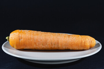Orange carrot on a plate isolated on a black background in a studio shot, unpeeled, a single carrot, ready to be prepared.