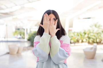 young pretty woman covering face with hands, peeking between fingers with surprised expression and looking to the side