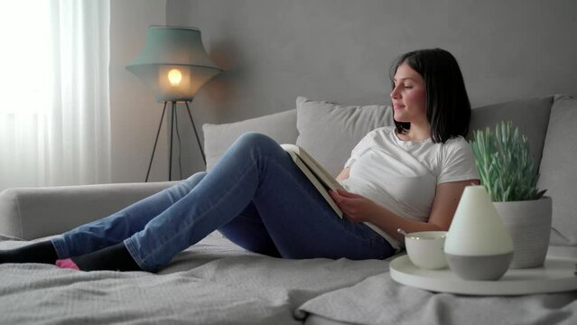 Beautiful dark hair young woman laying on cozy sofa and reading a book, essential oils diffusor next to her