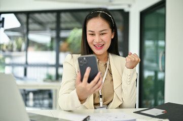 Obraz premium Excited Asian businesswoman showing clenched fist, looking at her smartphone screen