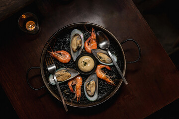 Delicious black risotto with shrimps and mussels on dinner wooden table, top view.