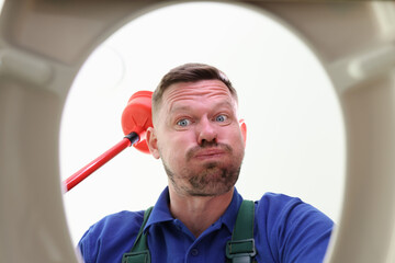 Unshaven man looks into toilet bowl holding red plunger