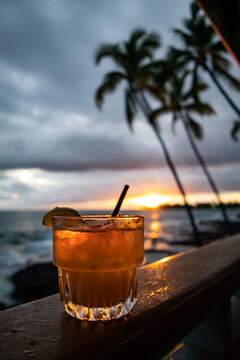 Photo of a mai tai cocktail on a wooden railing with the cloudy sunset and palm trees with ocean in the background on Big Island, Hawaii.