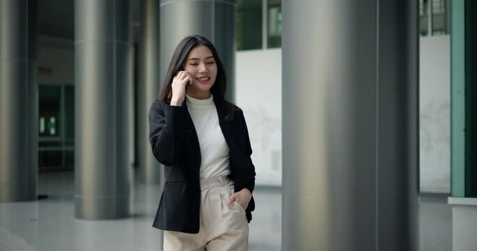 Front view shot, Footage of Young smiling elegant Asian busy business woman in a suit talking with a smartphone and walking in front of a modern business building. Business and people concepts.
