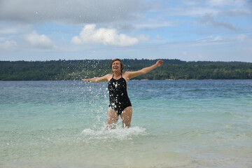 Happy mature woman 50-60 splashing in the water on the beach. Senior woman wear black swimsuit. Concept - travel, retirement, active elderly people, Philippines.