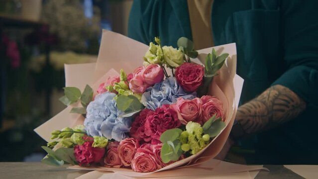 Close up shot of professional florist wrapping beautiful bouquet with wrapping paper. Vases of flowers at background. Process of working in flower shop. Floral business and entrepreneurship concept.