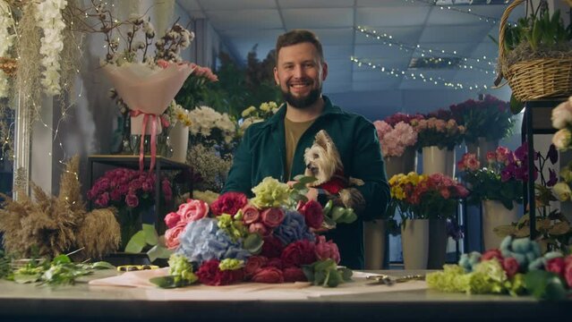 Professional male florist keeps cute dog in hands, smiles and looks at camera. Beautiful bouquet lies on table. Vases with flowers behind man. Concept of retail floral business and entrepreneurship.