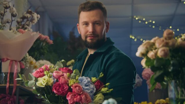 Professional florist collects and keeps beautiful bouquet in hands, smiles and looks at camera. Vases with fresh flowers stand behind man. Retail floral business and entrepreneurship concept. Portrait
