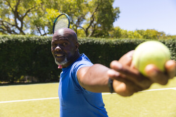 Happy senior african american man playing tennis, holding ball before serving on sunny grass court - Powered by Adobe