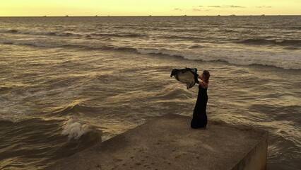 Elegant woman with scarf dancing on pier. Drone view. Sea with waves. Glamorous lady in black long dress.