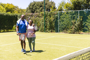 Happy senior african american couple with tennis rackets embracing on sunny grass court, copy space