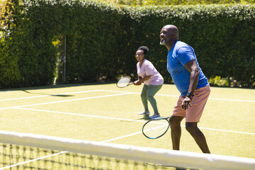 Happy senior african american couple playing tennis doubles on sunny grass tennis court, copy space - Powered by Adobe