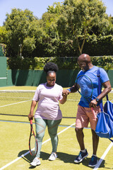 Obraz premium Happy senior african american couple with rackets holding hands on sunny tennis court, copy space
