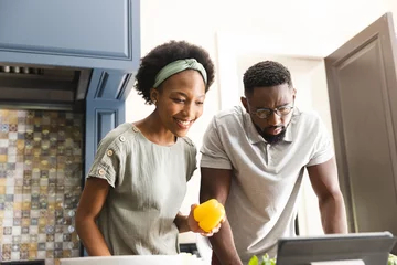 Tuinposter Happy african american couple preparing meal together using tablet in kitchen © wavebreak3