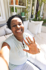 Portrait of smiling senior african american woman waving during video call in sunny living room