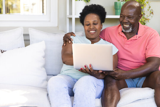 Happy senior african american couple on couch using laptop and embracing in sunny living room