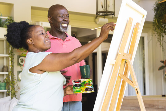 Happy senior african american couple painting canvas on easel together at home