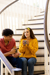 Biracial young couple holding coffee cups and talking while sitting on staircase at home, copy space