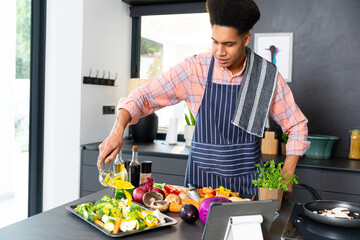 Happy biracial man wearing apron cooking dinner, pouring vegetables in kitchen, unaltered