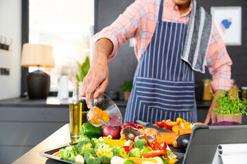 Midsection of biracial man wearing apron cooking dinner, pouring vegetables in kitchen