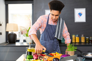 Biracial man wearing apron cooking dinner, pouring vegetables with oil in kitchen, unaltered