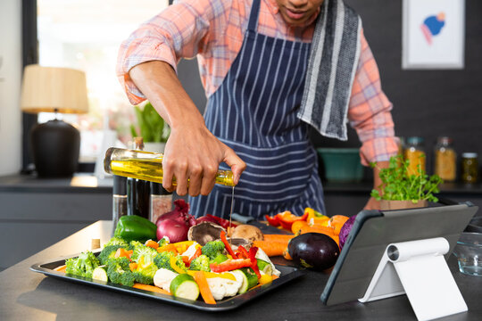 Biracial man wearing apron cooking dinner, pouring vegetables with oil in kitchen, unaltered