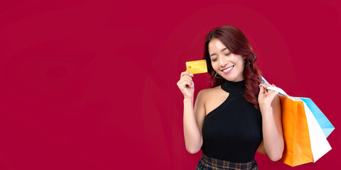 Banner: Stylish Asian woman in black dress holding shopping bags holding gold credit cards on crimson background. Beautiful Thai woman holding shopping bags holding gold credit cards on red background