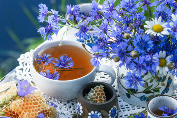Cornflower herbal tea in white cup on white crochet napkin on table outdoors, healthy cornflower drink with honey in nature background, fresh cornflowers flowers in vase, healthcare and healthy eating