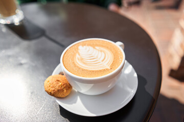 A cup of hot cappuccino with cookies on the table in the coworking space of the coffee shop. A cup of coffee on the veranda on a sunny day. Coffee break at the cafe. Close-up