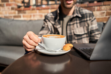 A young man is sitting on the veranda in a cafe working and drinking coffee Handsome successful smart man looking at a laptop. Reading, browsing the Internet