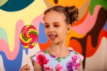 Joyful little girl stuck out her tongue, looking on round lollipop at multi colored wall. Lovely...