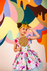 Cute little girl in flowers dress holding round giant lollipop at colored wall, looking at camera. Child 6-7 year old in pineapple sunglasses with candy on stick. Summer fashion concept. Copy ad space