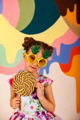 Adorable little girl in flowers dress holding round giant lollipop at colored wall, looking at camera. Kid 6-7 year old in pineapple sunglasses with candy on stick. Summer fashion concept. Copy space