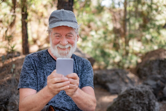 Portrait of smiling senior bearded man sitting in the forest during a mountain excursion looking at mobile phone. Elderly active grandfather resting using smartphone