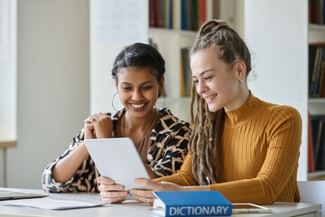 Two smiling girls watching online lesson on tablet pc while sitting at table in library