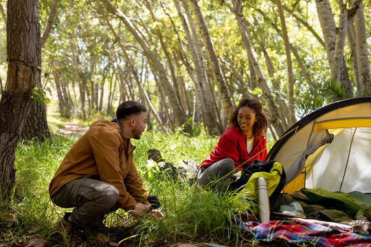 Happy biracial couple pitching tent together in forest, unaltered
