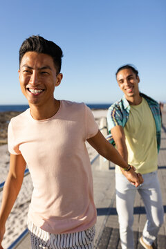 Happy biracial gay male couple holding hands on sunny promenade by the sea