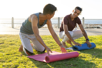 Happy biracial gay male couple rolling yoga mats on promenade by the sea