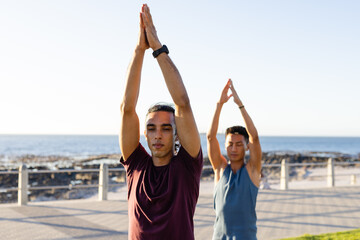 Happy biracial gay male couple practicing yoga and meditating on promenade by the sea