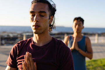 Biracial gay male couple practicing yoga and meditating on promenade by the sea
