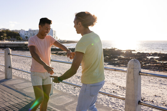 Happy biracial gay male couple having fun dancing on promenade by the sea