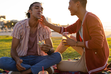 Happy biracial gay male couple having picnic on promenade by the sea at sundown